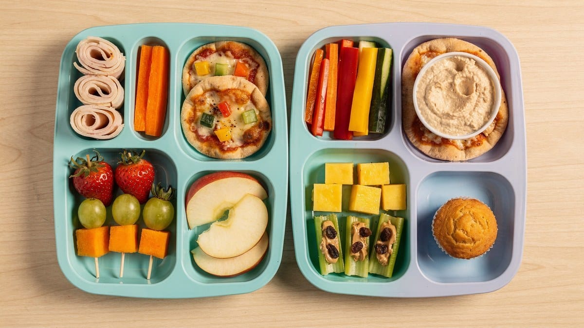 Two bento lunch boxes with mini pizzas, turkey roll-ups, veggie sticks, hummus, fruit skewers, apple slices and a muffin
